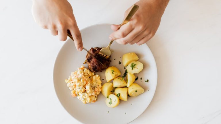 Top view of crop unrecognizable male with cutleries slicing delicious beef cutlet garnished with boiled potatoes and parsley and portion of Russian meat salad on plate placed on white table